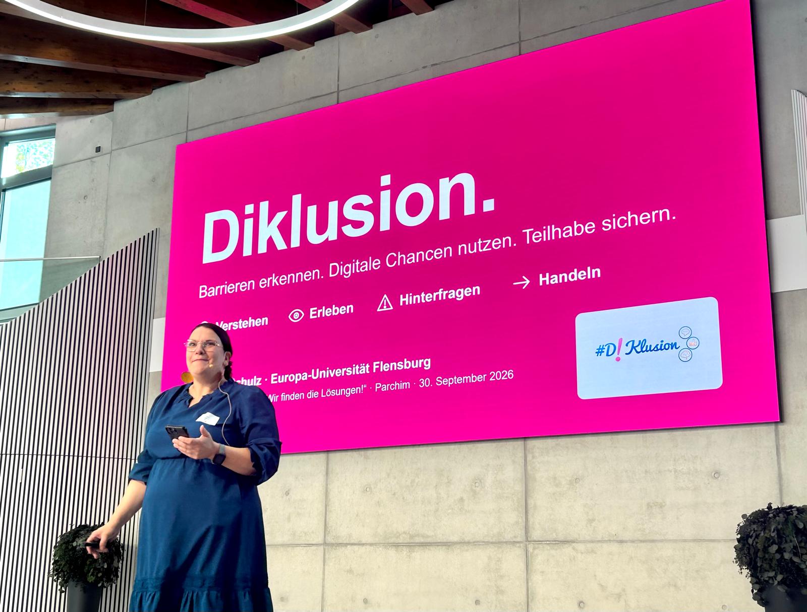 Dr. Lea Schulz spricht auf einer Bühne, hinter ihr eine magentafarbene Folie mit dem Titel „Diklusion. Barrieren erkennen. Digitale Chancen nutzen. Teilhabe sichern.“ und dem Logo #Diklusion