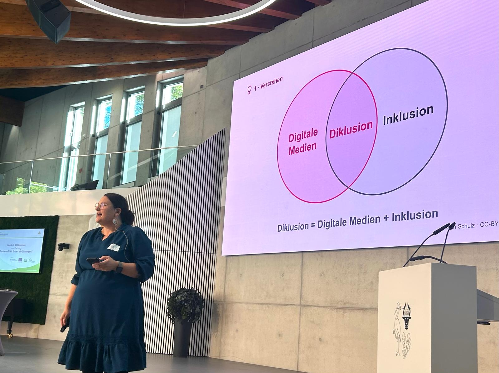Dr. Lea Schulz auf einer Bühne, hinter ihr eine Folie mit zwei sich überschneidenden Kreisen „Digitale Medien“ und „Inklusion“, die Schnittmenge heißt „Diklusion“, darunter die Formel Diklusion = Digitale Medien + Inklusion